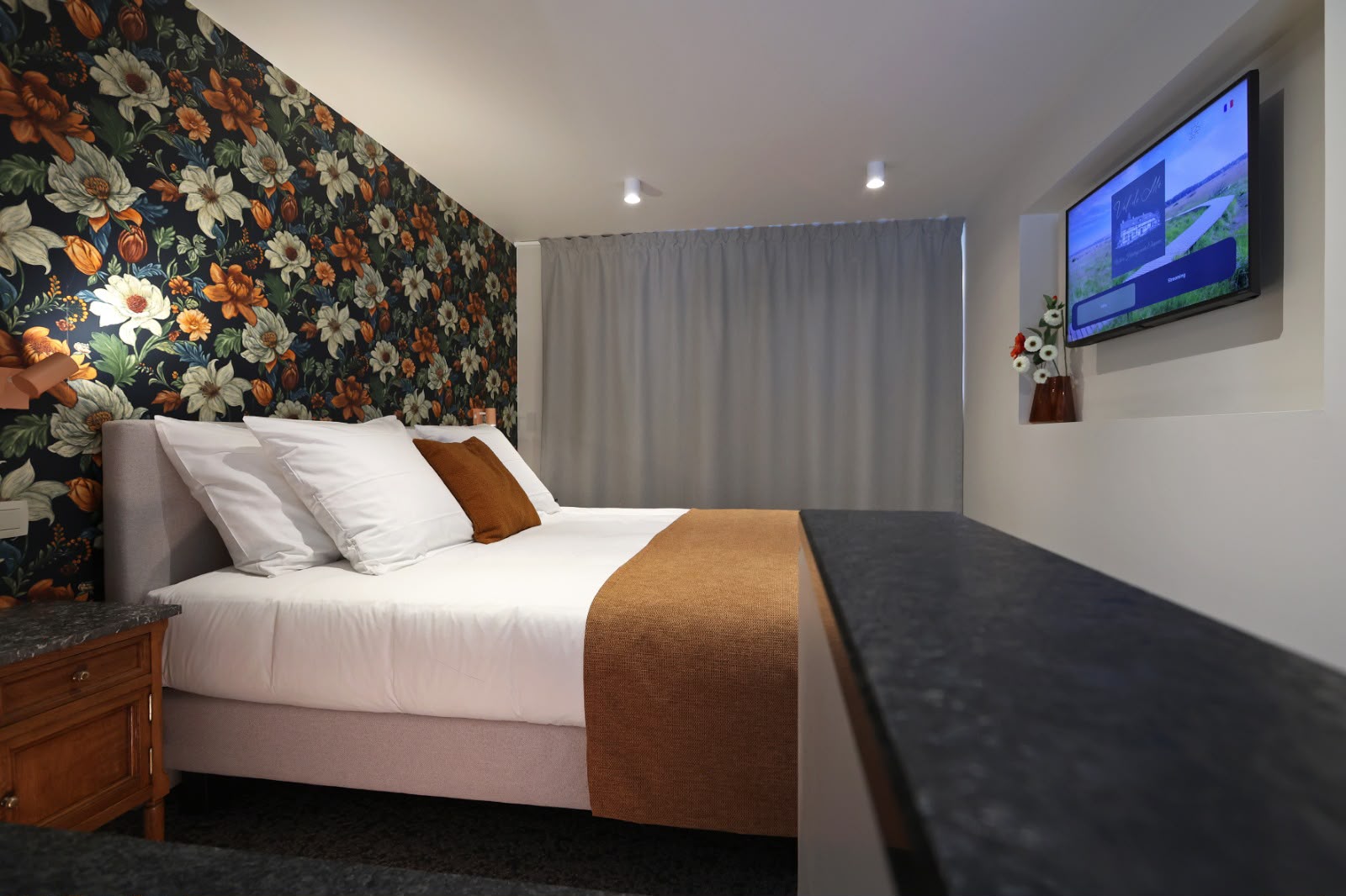 L'Ambre : lit sous un papier peint à grandes fleurs cuivrées, paroi vitrée ouvrant sur la salle de bain